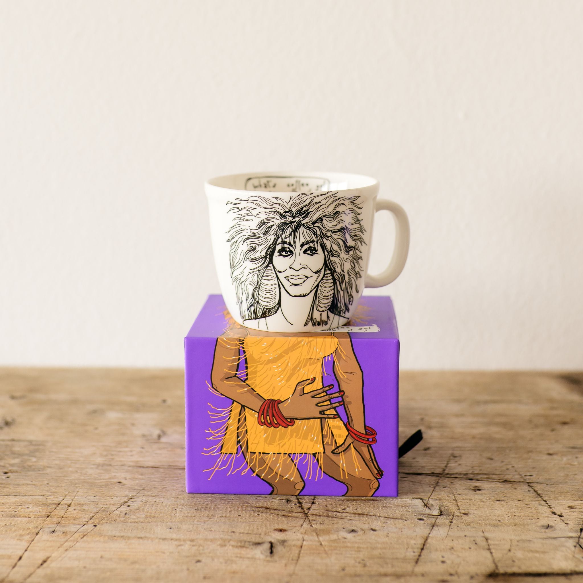 Mug with artistic design inspired by tina turner,wearing a yellow dress with fringe on a purple box on a wooden surface.