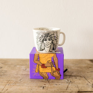 Mug with artistic design inspired by tina turner,wearing a yellow dress with fringe on a purple box on a wooden surface.